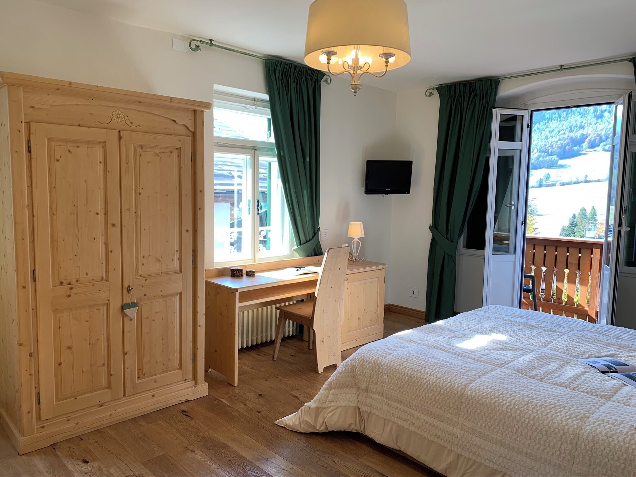 A bedroom with a wooden wardrobe, a desk, a bed with a white duvet, a chandelier, a TV, and a balcony with a view of the outdoors.