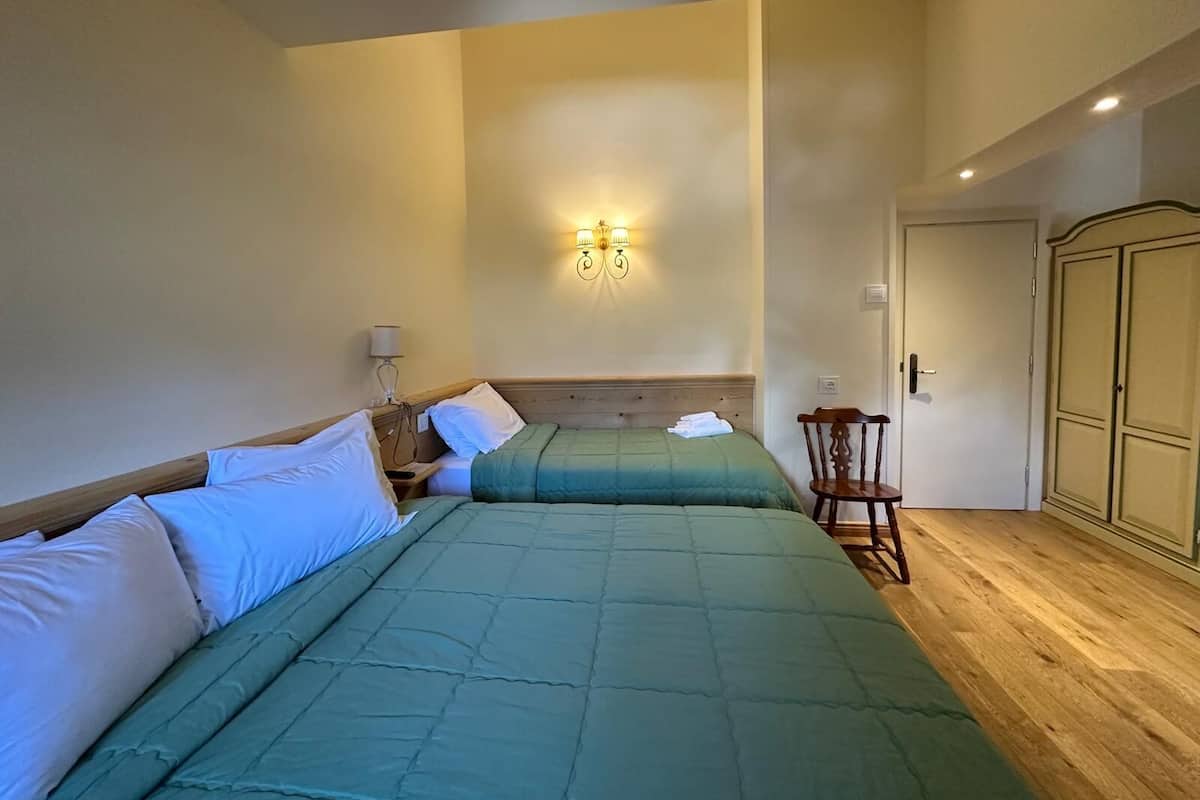 A hotel room with two beds, a wooden wardrobe, a chair, and a wall-mounted light fixture.
