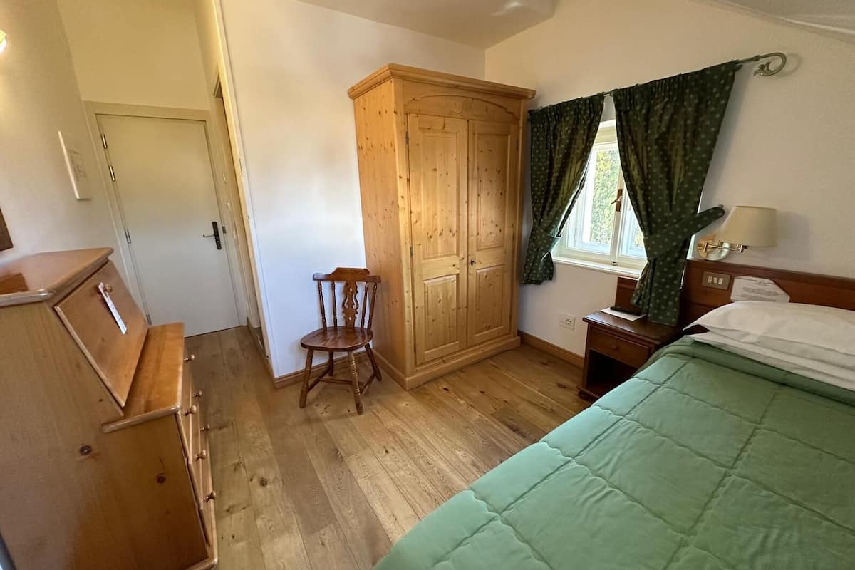 A bedroom with a wooden wardrobe, a chair, a bed with green bedding, a window with curtains, and a bedside table with a lamp.