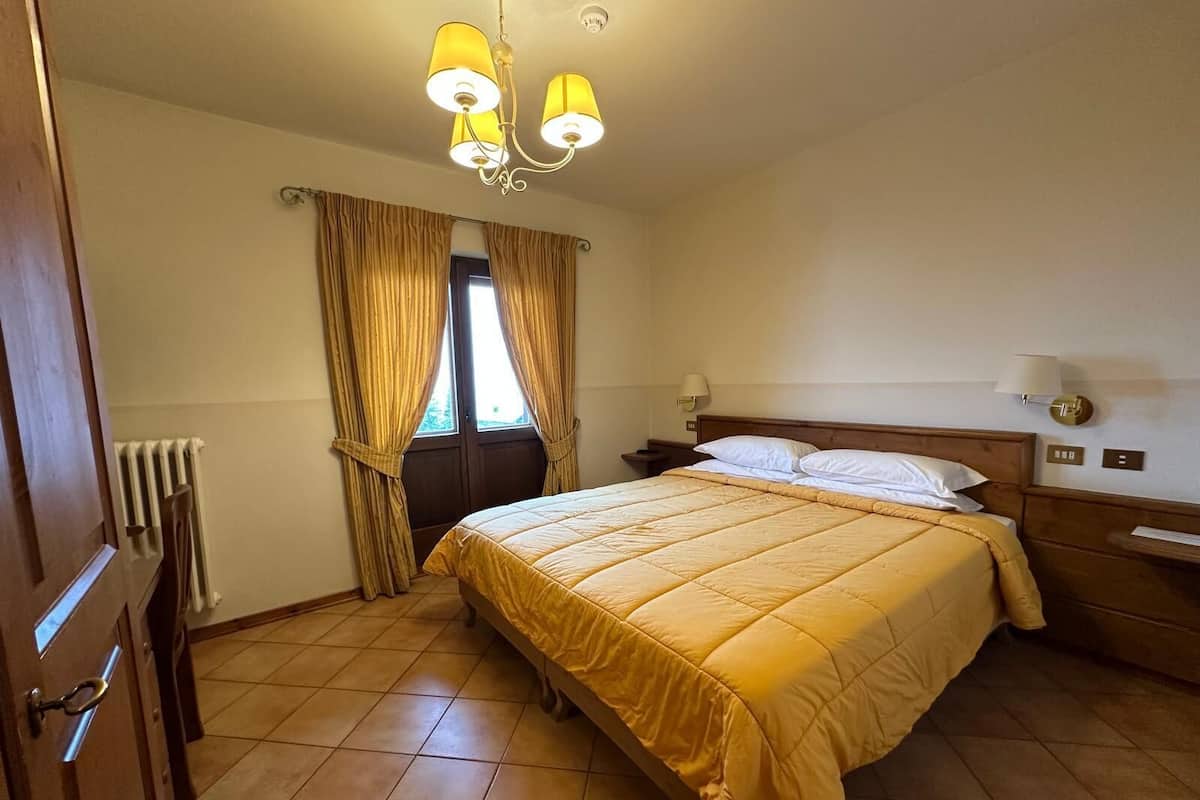A hotel room with a bed, a nightstand, a lamp, a window with curtains, and a radiator.