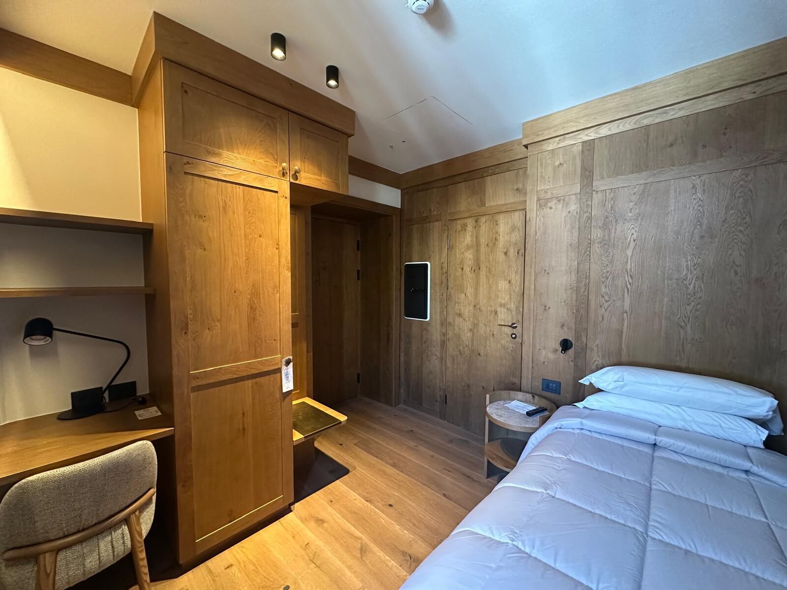 Un dormitorio con un armario de madera, un escritorio con una silla, una cama con ropa de cama blanca y una mesita de noche.