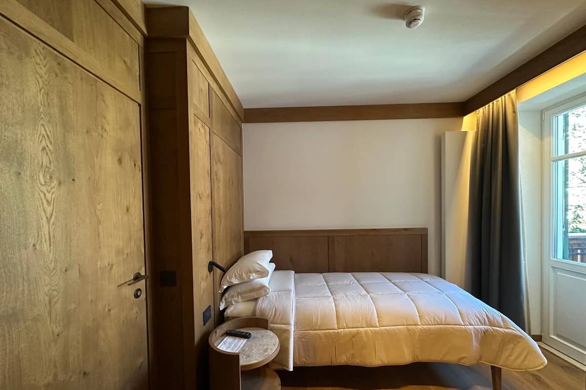 A bedroom with a wooden wardrobe, a round bedside table, a bed with a headboard, and a window with curtains.