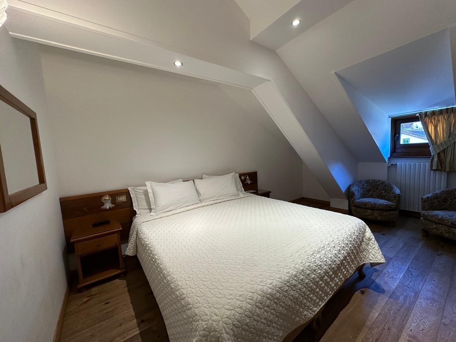 A bedroom with a sloped ceiling, a bed with white bedding, a nightstand, a chair, and a window with curtains.
