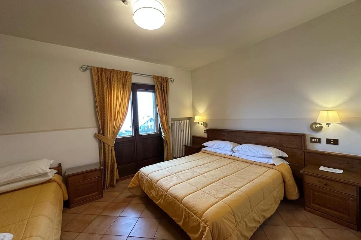 A hotel room with a large bed, a smaller bed, a window with curtains, a radiator, and wall-mounted lamps.