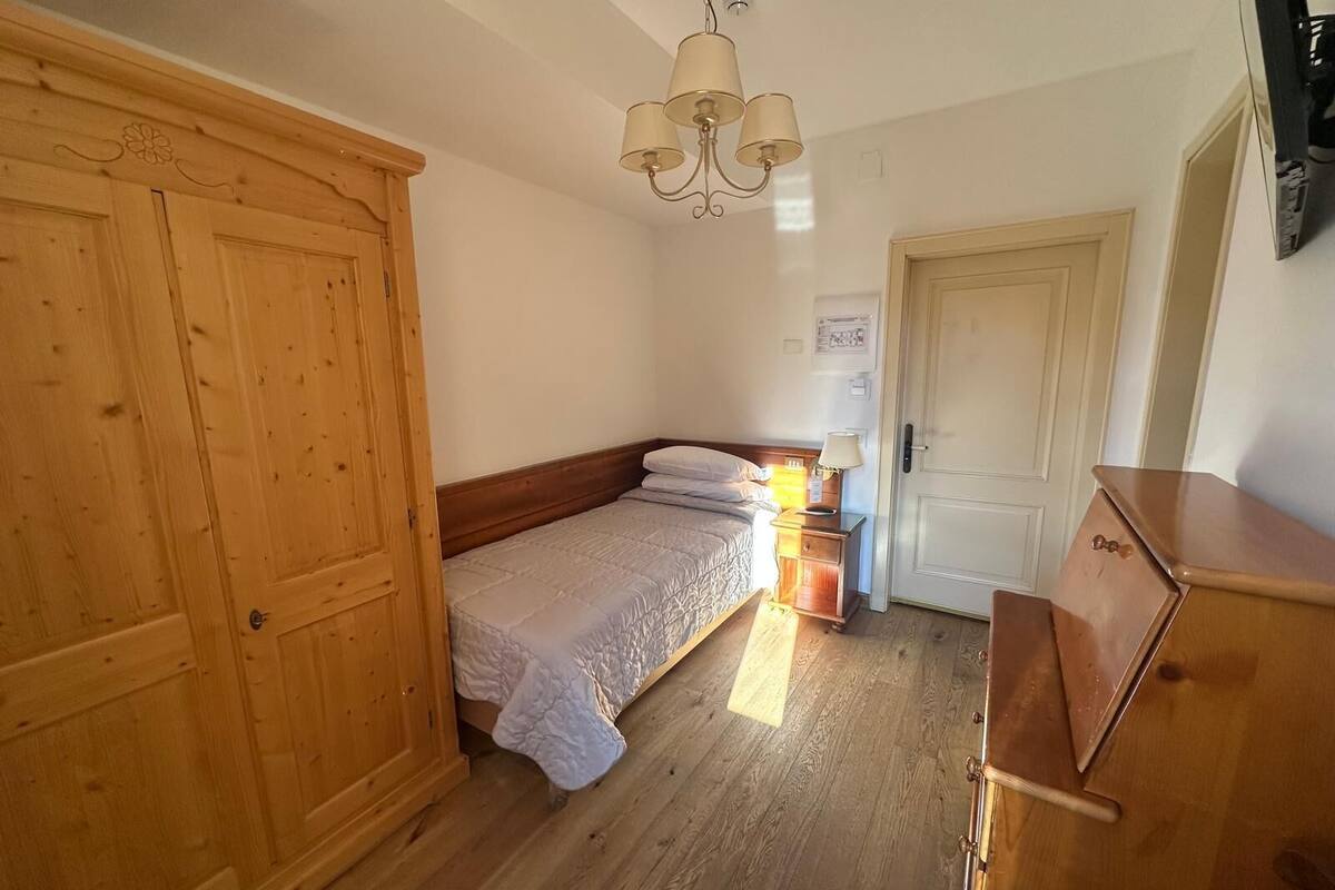 A bedroom with a wooden wardrobe, a bed with a white duvet, a bedside table with a lamp, and a wooden dresser.