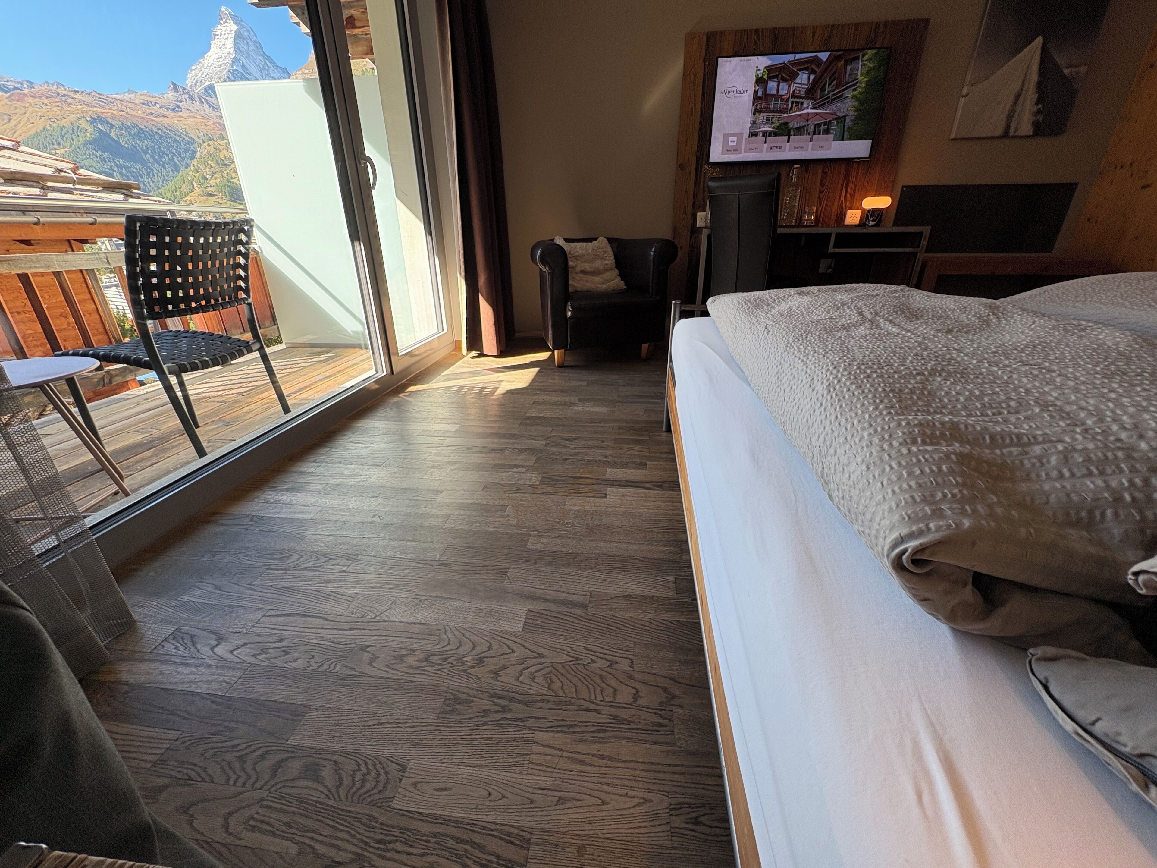 Habitación de hotel con una cama grande, un televisor montado en la pared, piso de madera, una silla y vistas a las montañas a través de una puerta corredera.