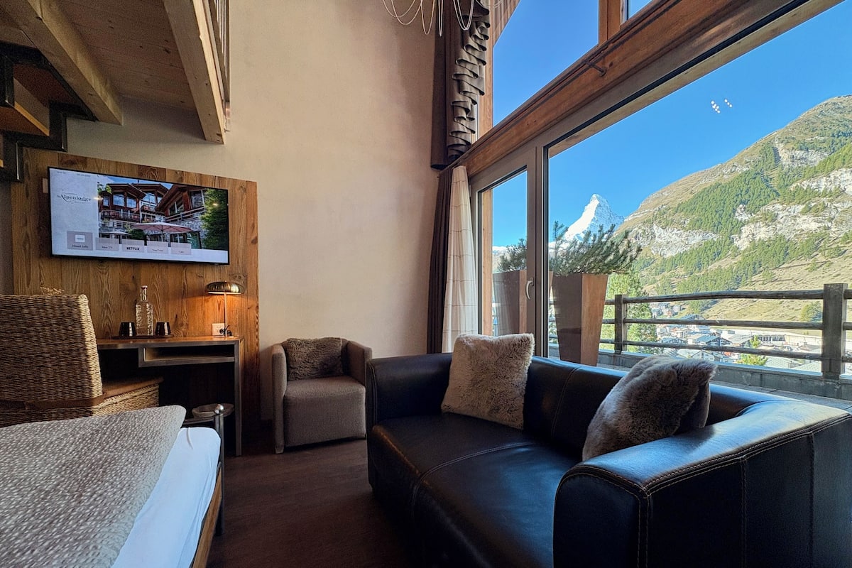 Une pièce confortable avec un lit, un canapé en cuir, un petit fauteuil, un téléviseur fixé au mur et une grande fenêtre donnant sur les montagnes.