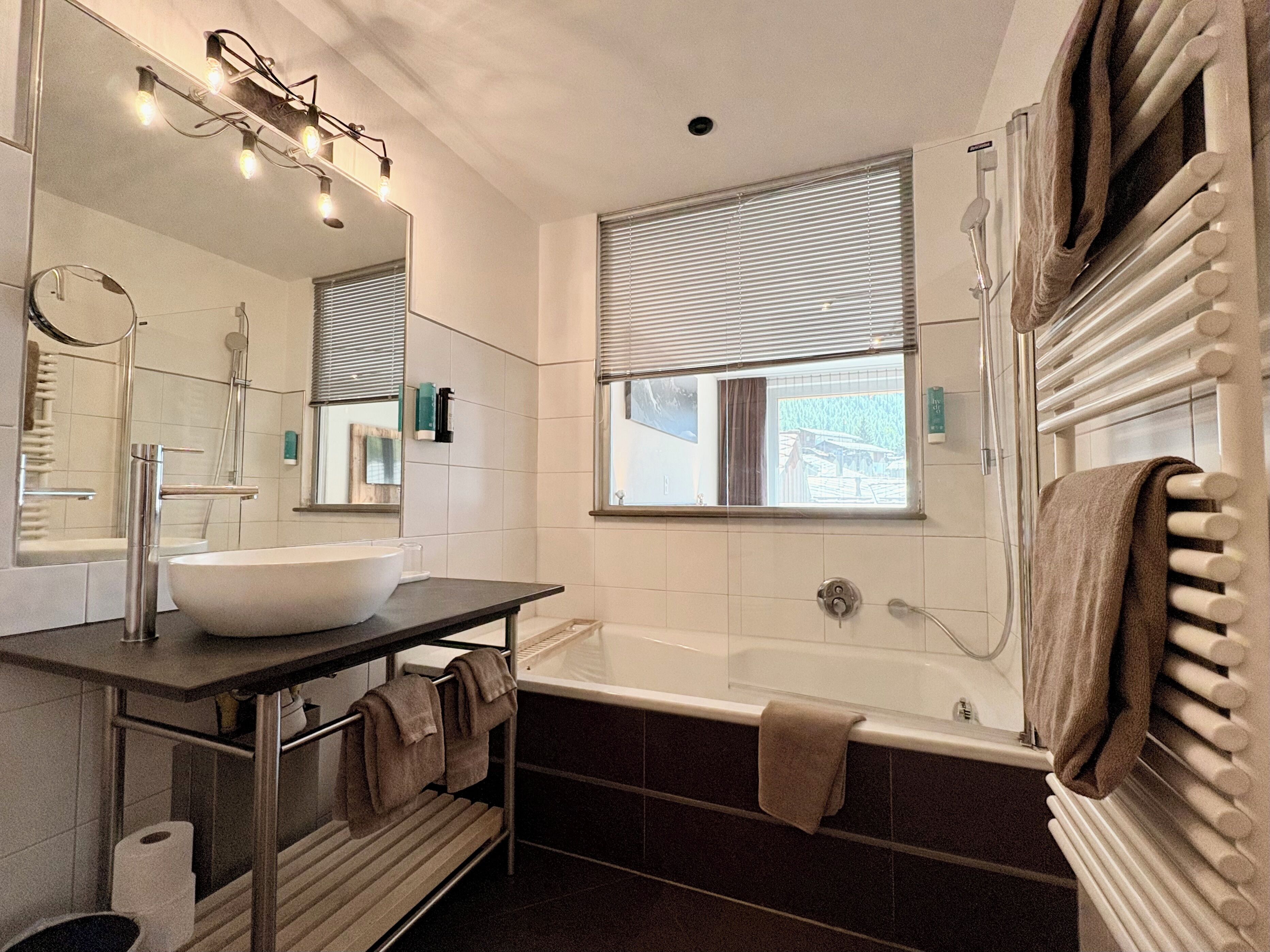 Un bagno moderno con una vasca da bagno ampia, un mobile lavabo con lavandino, uno specchio e una finestra con persiane.