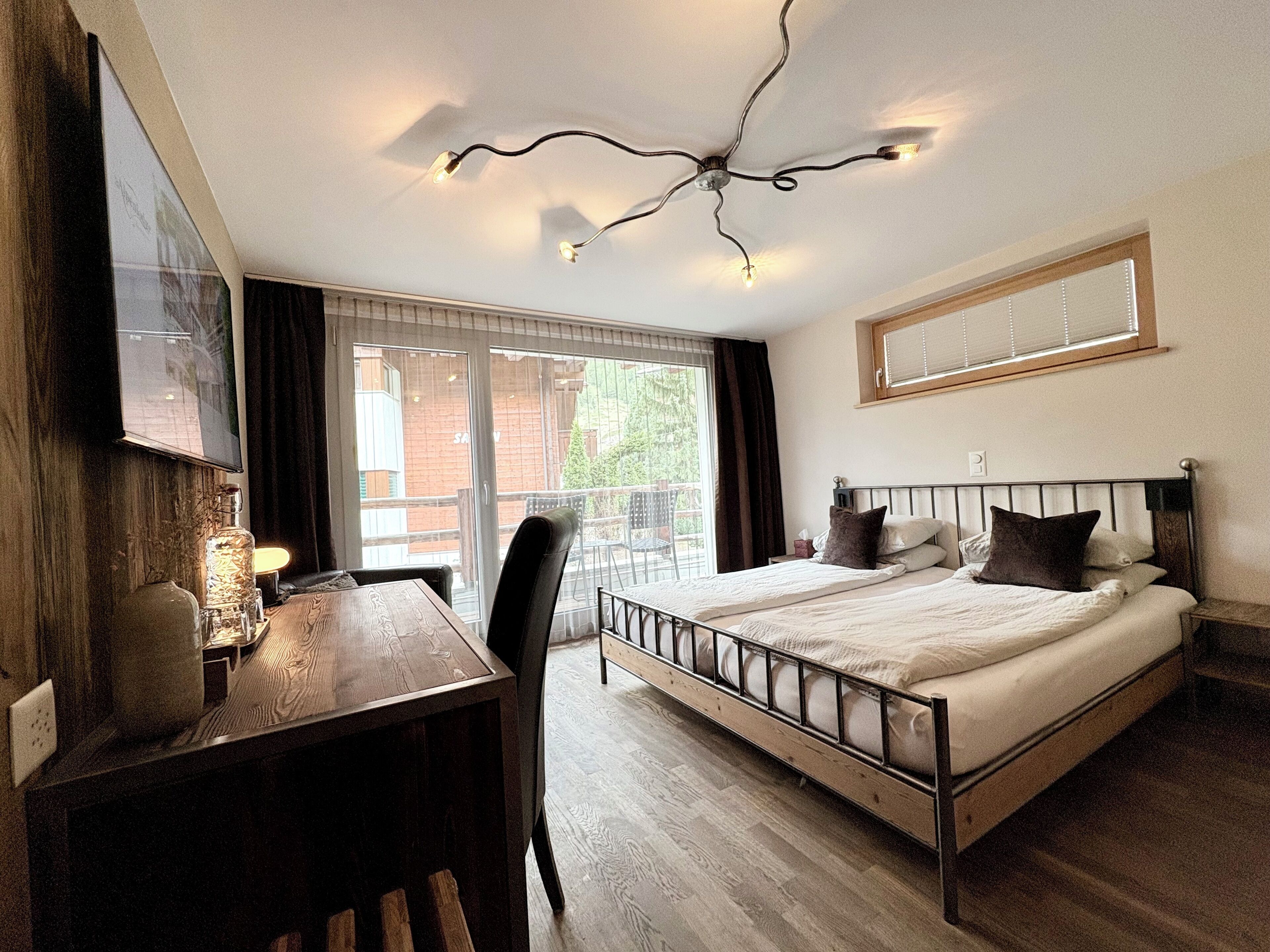 A bedroom with a bed, a desk, and a chair. There is a window with blinds and a view of the outside.