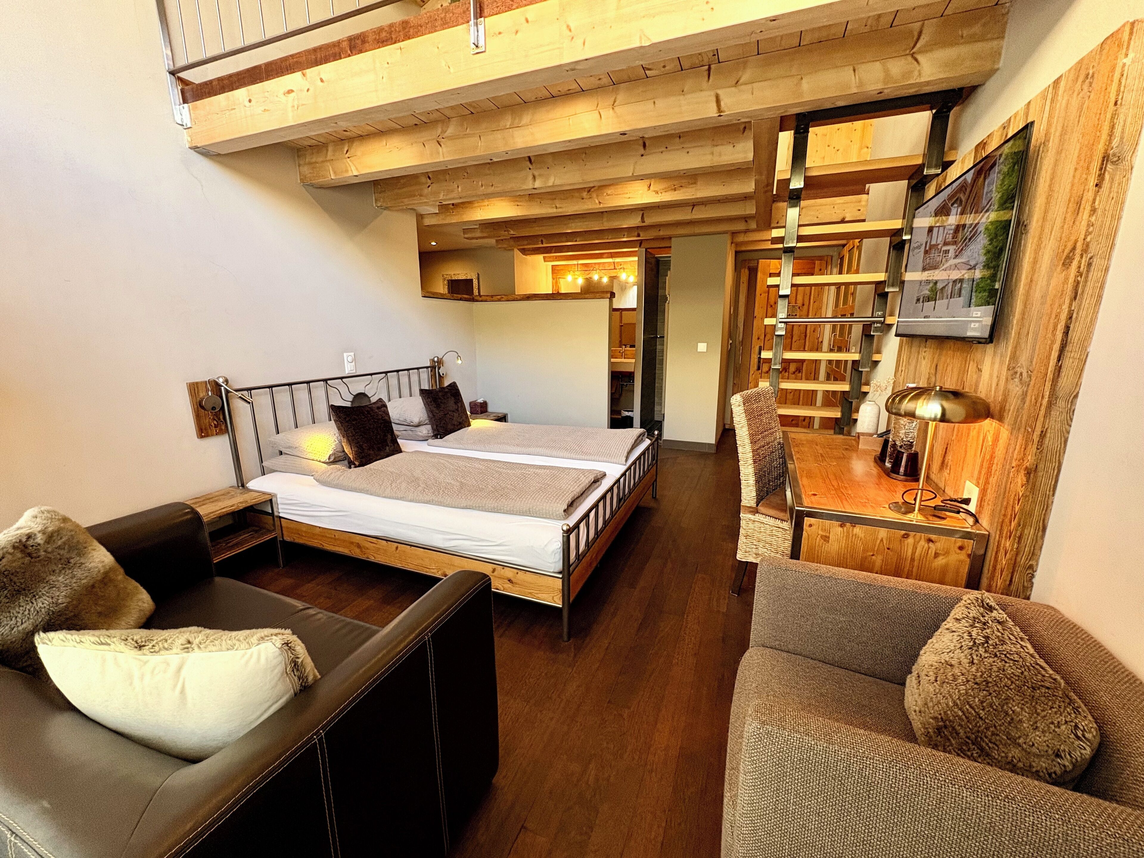 Een gezellige slaapkamer met een houten plafond, een bed met kussens, een tafeltje met een lamp en een stoel.
