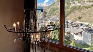 A chandelier with multiple light bulbs and hanging crystals, a wooden ceiling, and a large window with a view of a mountain and a town. - Alpenlodge (Zermatt)