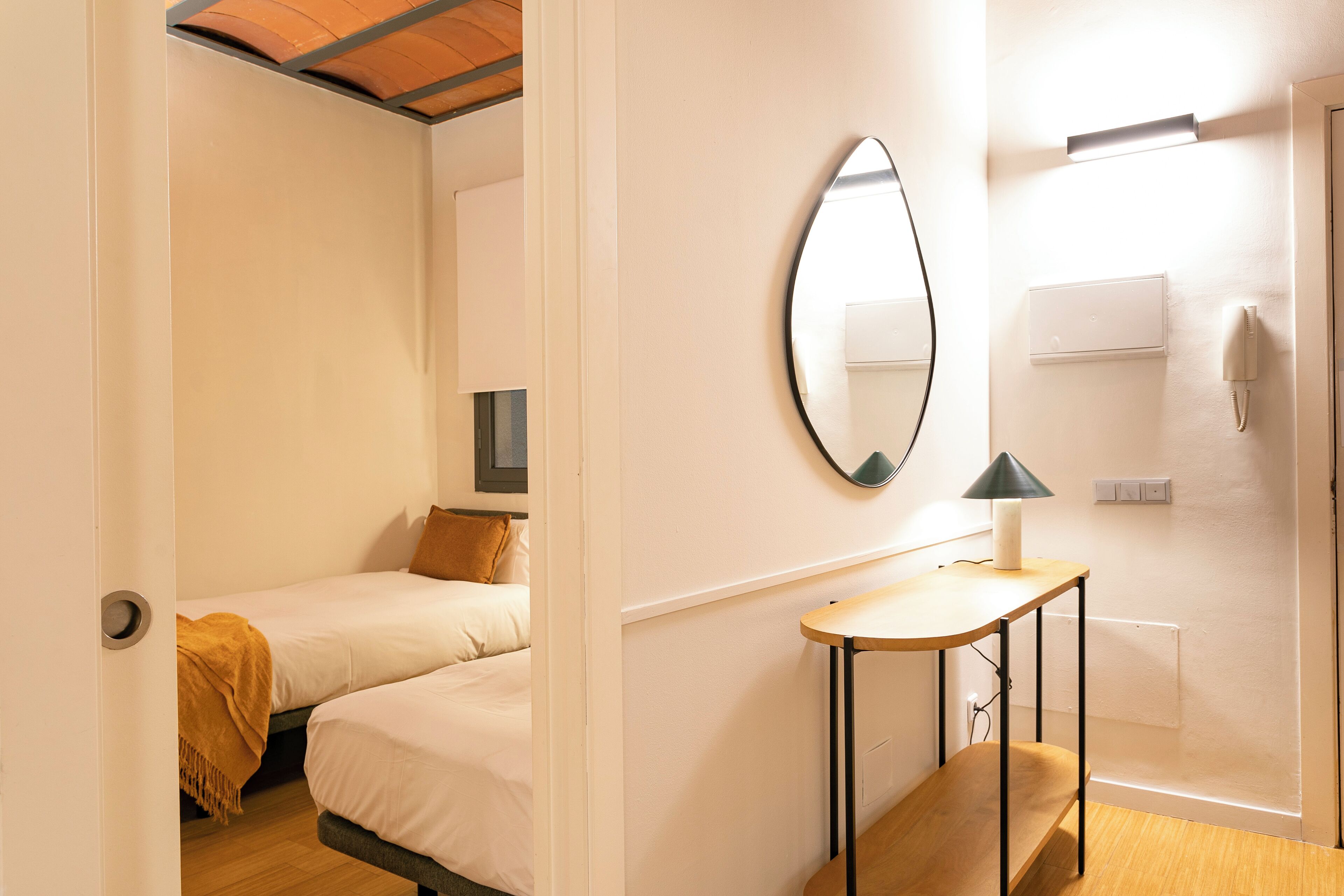 A hotel room with two beds, a bedside table with a lamp, a mirror, a wall-mounted phone, and a light switch.
