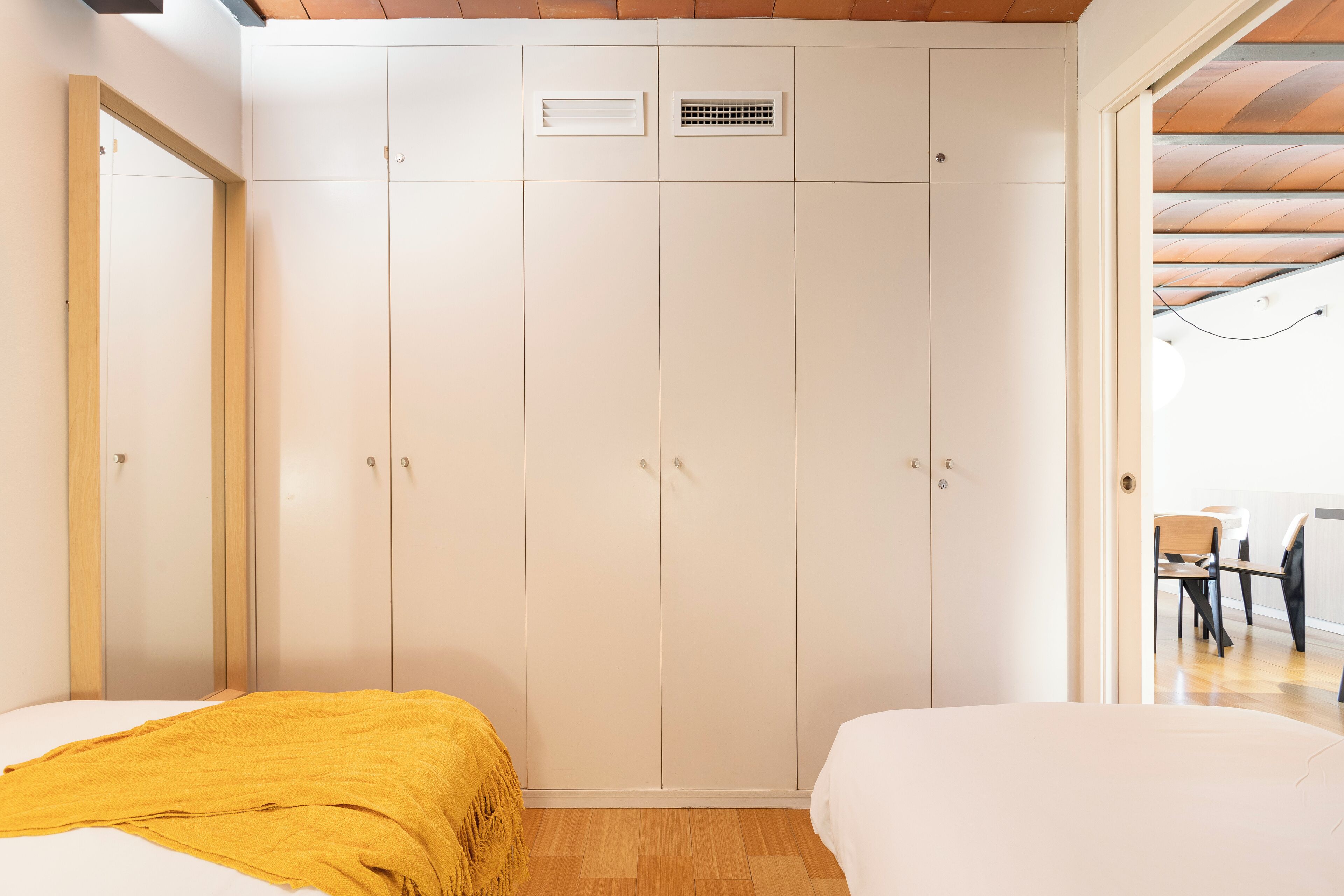 Une chambre avec une grande armoire, un lit recouvert d’une couverture jaune et une vue sur un coin repas, visible à travers une porte ouverte.