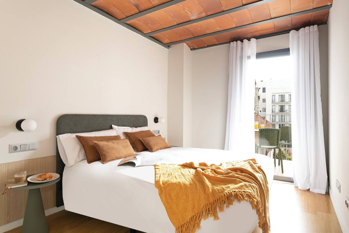 A modern hotel room with a large bed, a side table with a croissant, and a view of the city through the window.