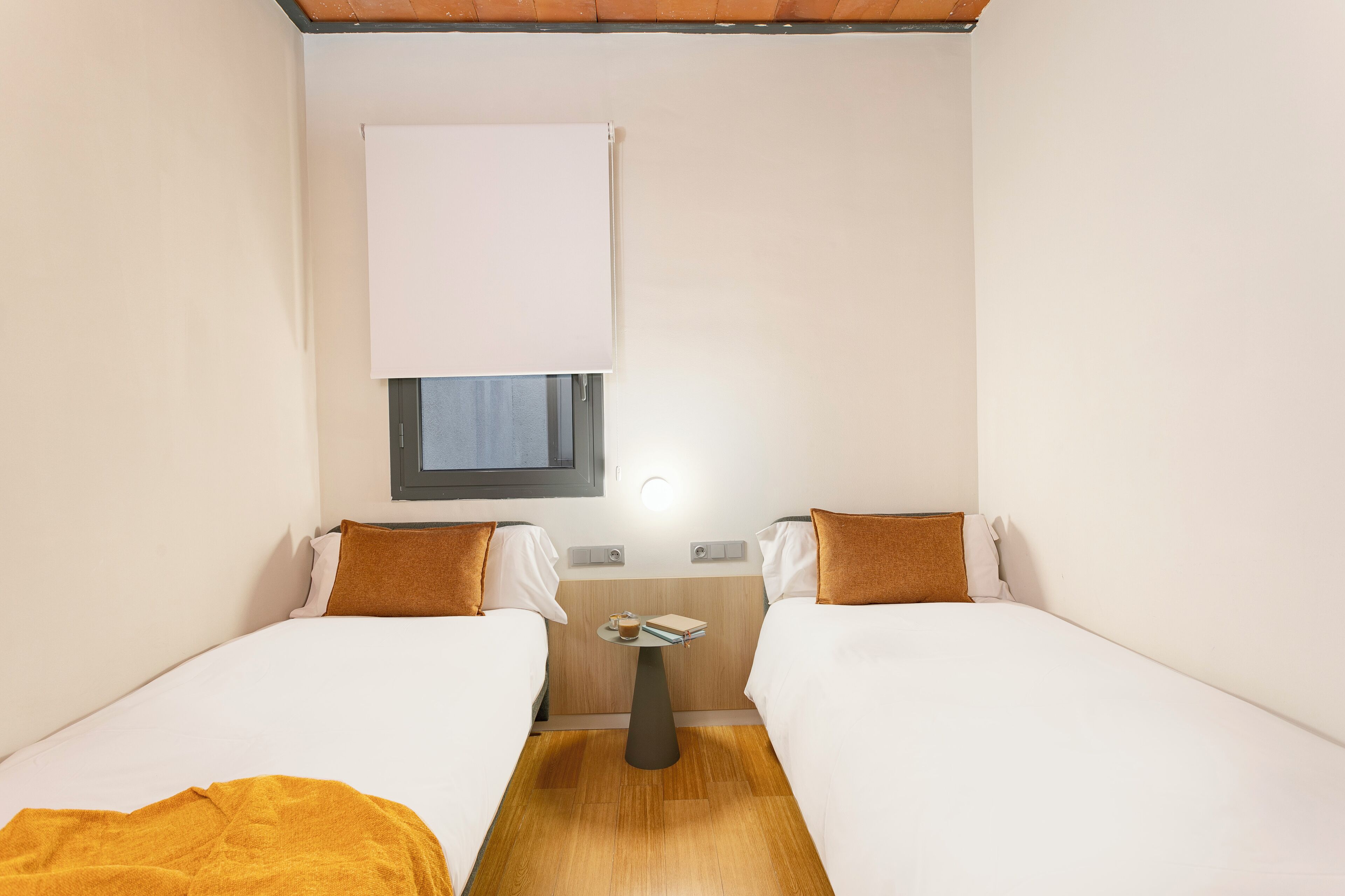 Een compacte, goed verlichte kamer met twee bedden, een tafeltje en een raam met een jaloezie.