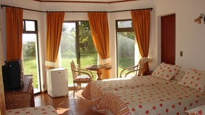 Una habitación con una cama, una mesita y una silla. Hay una ventana con cortinas, y se ve una puerta al fondo.