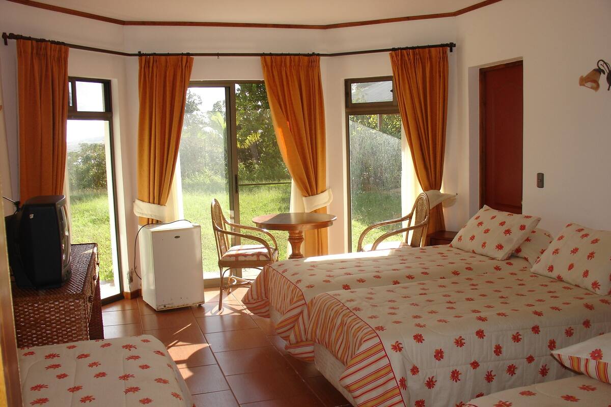 A room with a bed, a small table, and a chair. There is a window with curtains, and a door is visible in the background.