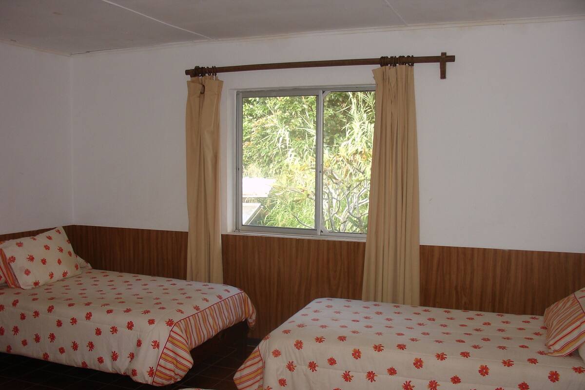 A room with two beds, each with floral-patterned bedding. There is a window with curtains, and the room has wooden paneling on the walls.