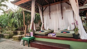 A covered outdoor seating area with two cushioned benches, a table with a green tablecloth, and potted plants. - Bounty Hotel (Kuta)