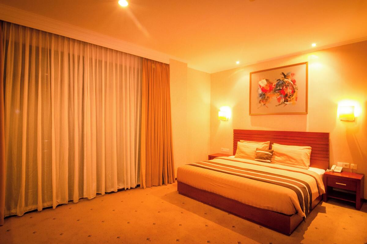 A hotel room with a large bed, a nightstand, a wall-mounted painting, and a window with curtains.
