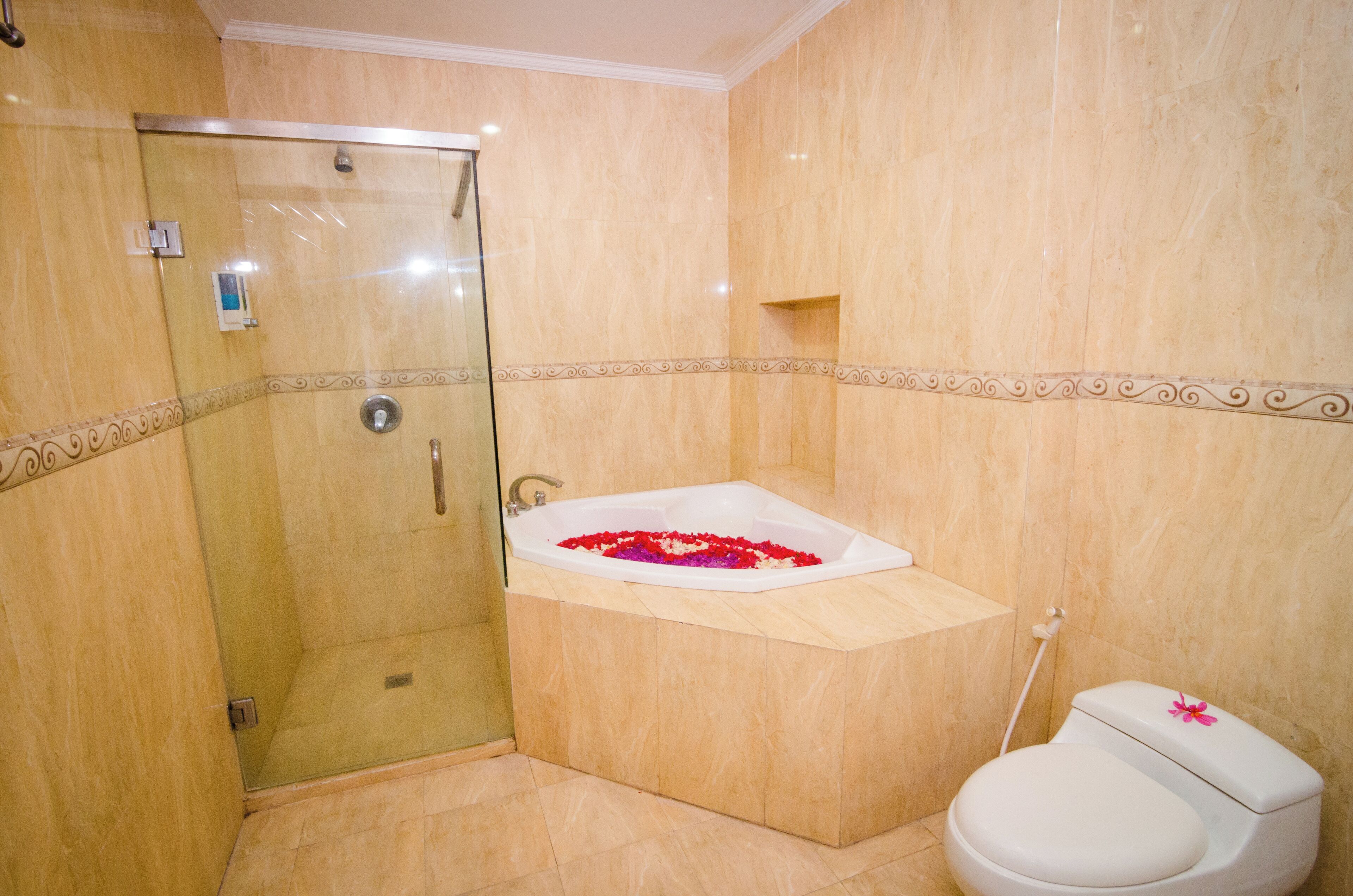 Casa de banho com chuveiro em cabine de vidro, banheira com pétalas de rosa e sanita decorada com uma flor cor-de-rosa.