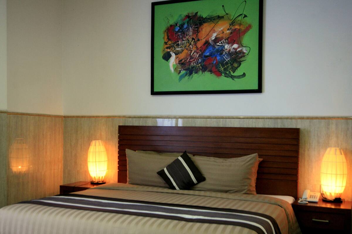 A hotel room with a bed, two bedside lamps, a telephone, and a framed abstract painting on the wall.