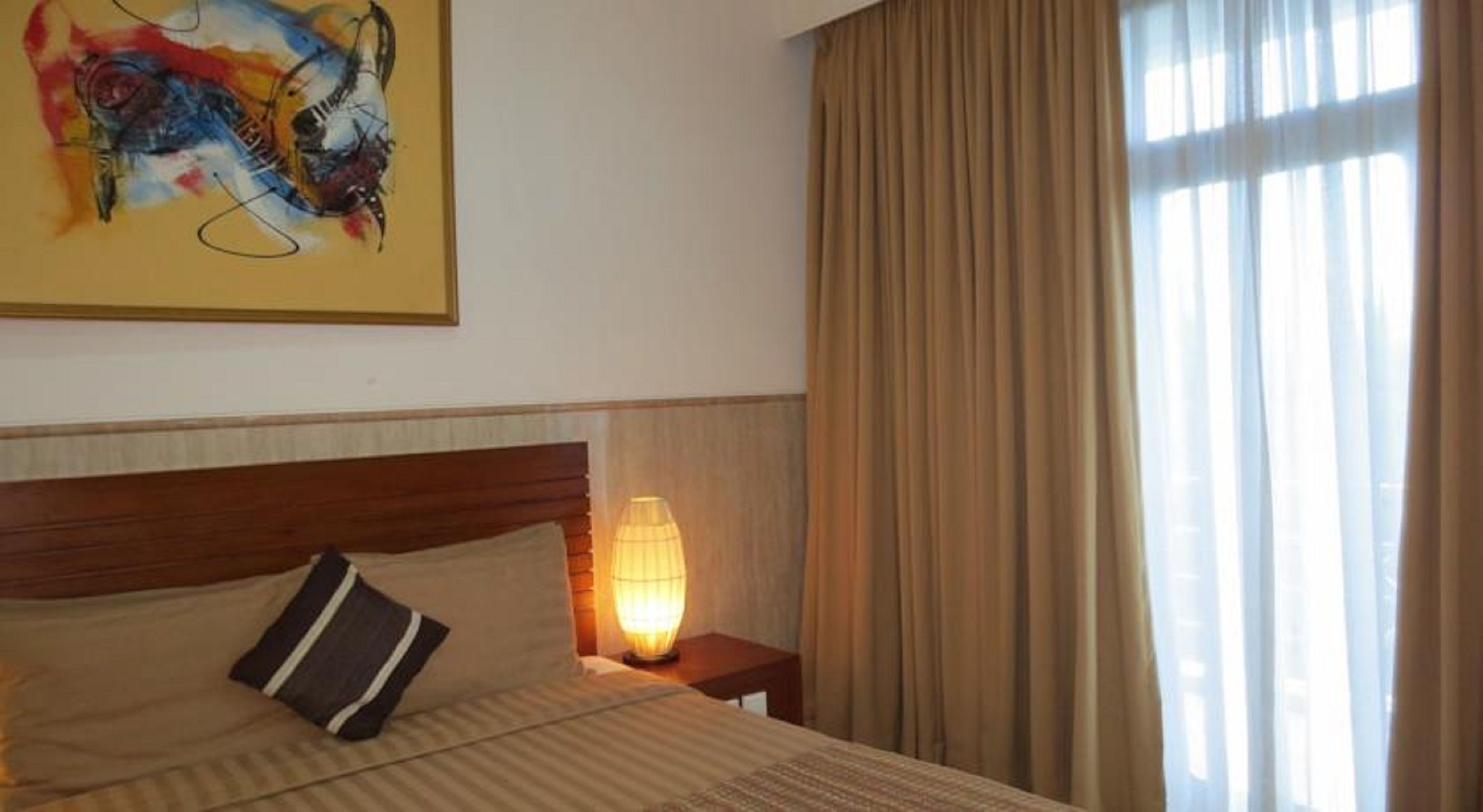 A hotel room with a bed, a nightstand, a lamp, a painting on the wall, and a window with curtains.