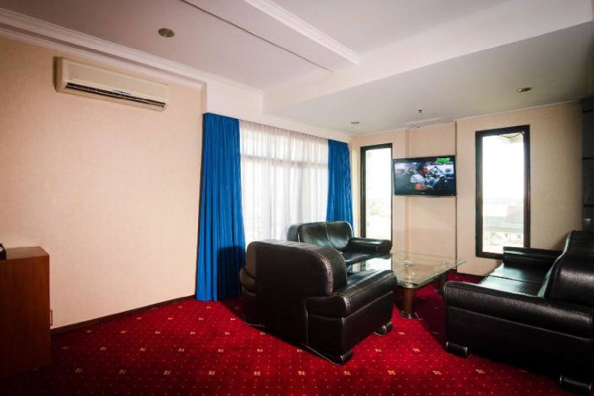 A hotel room with a red carpet, a leather sofa, a glass coffee table, and a TV on the wall.