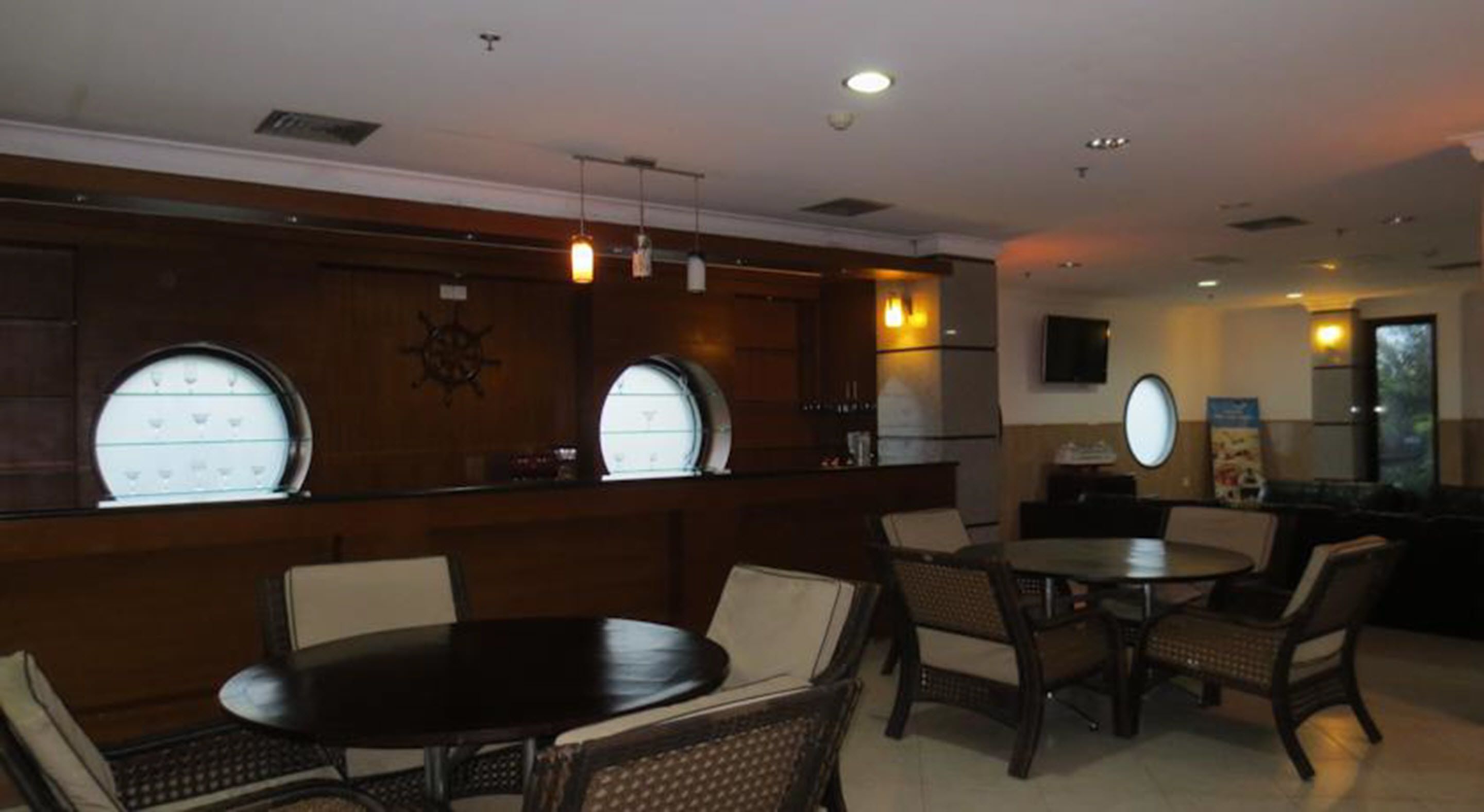 Un restaurante con mesas y sillas redondas, una zona de bar con decoración de timón de barco y un televisor montado en la pared.