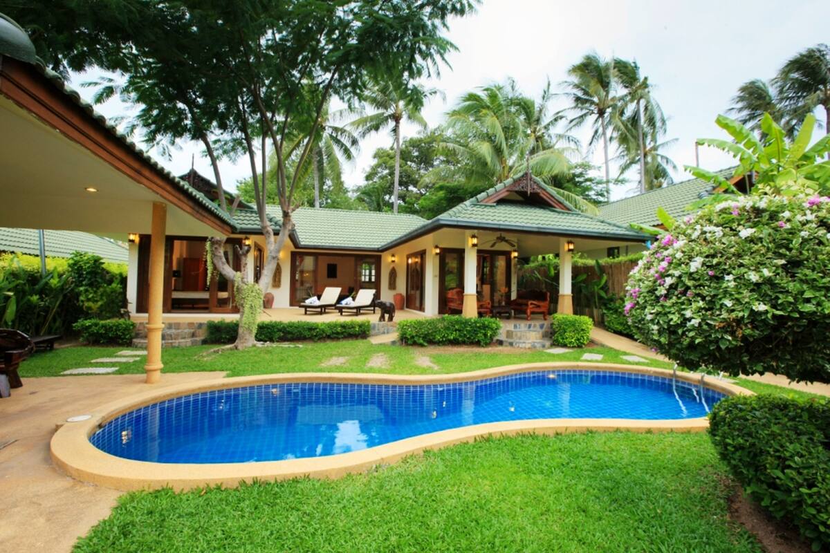 A poolside view of a resort with a swimming pool, lounge chairs, and tropical landscaping.