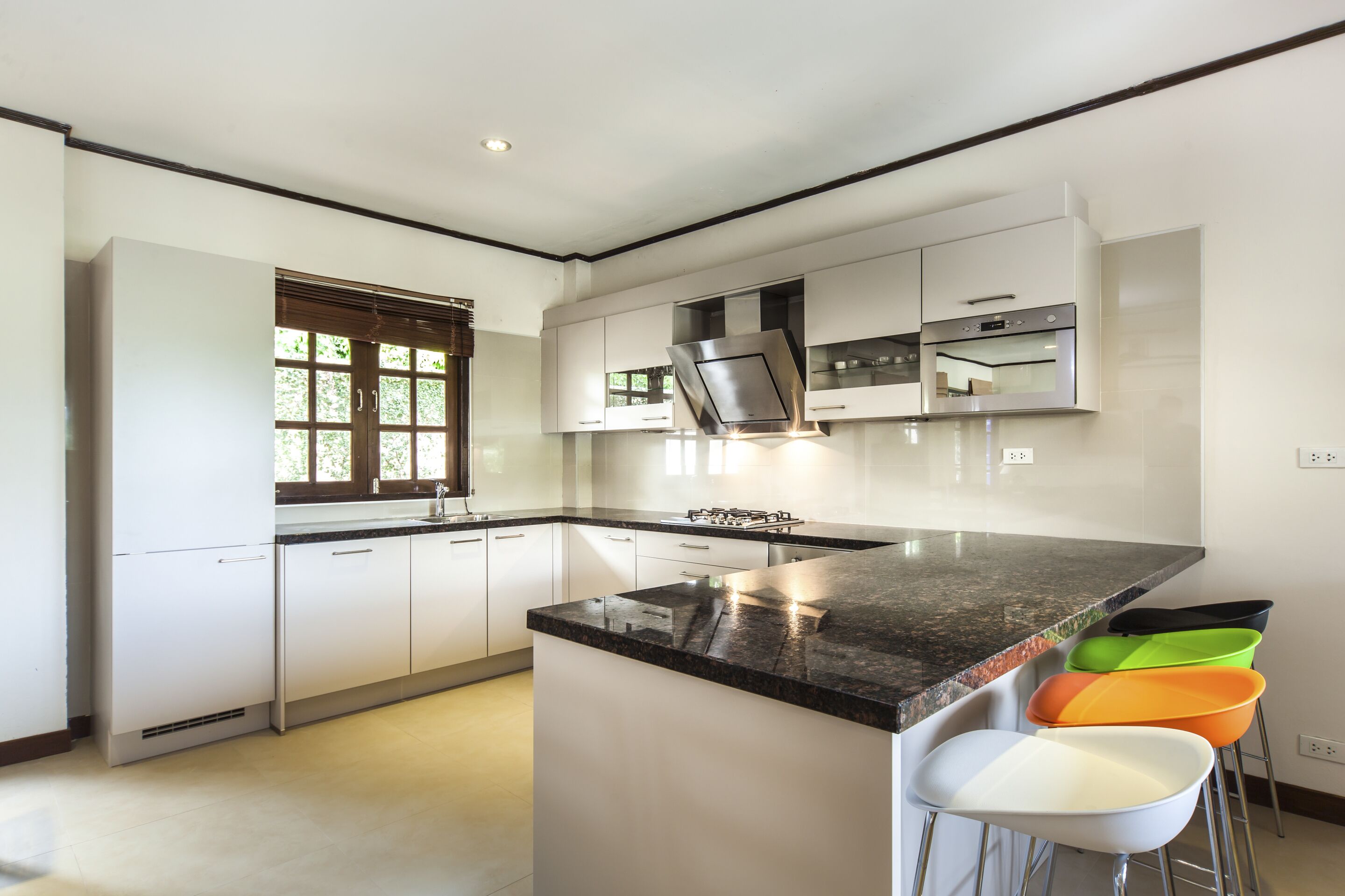 A modern kitchen with white cabinets, a black countertop, and a gas stove. There are three bar stools with colorful seats.