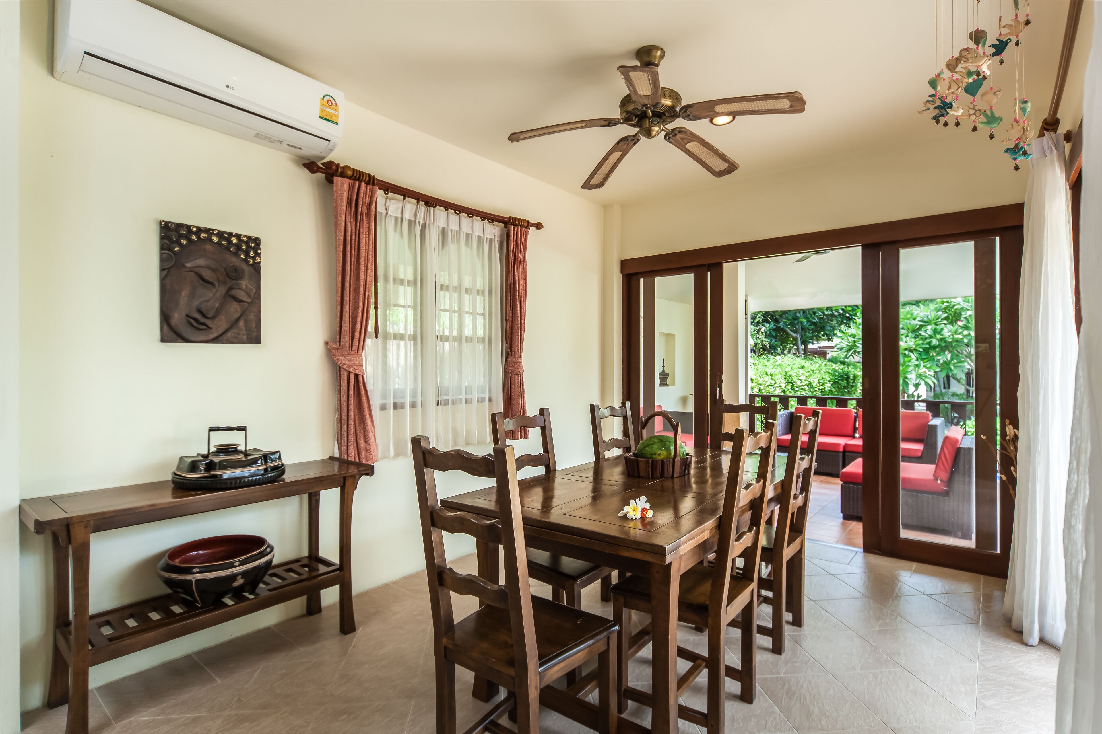 A dining area with a wooden table, chairs, and a ceiling fan. There is a window with curtains, a wall-mounted air conditioner, and a view of an outdoor seating area.