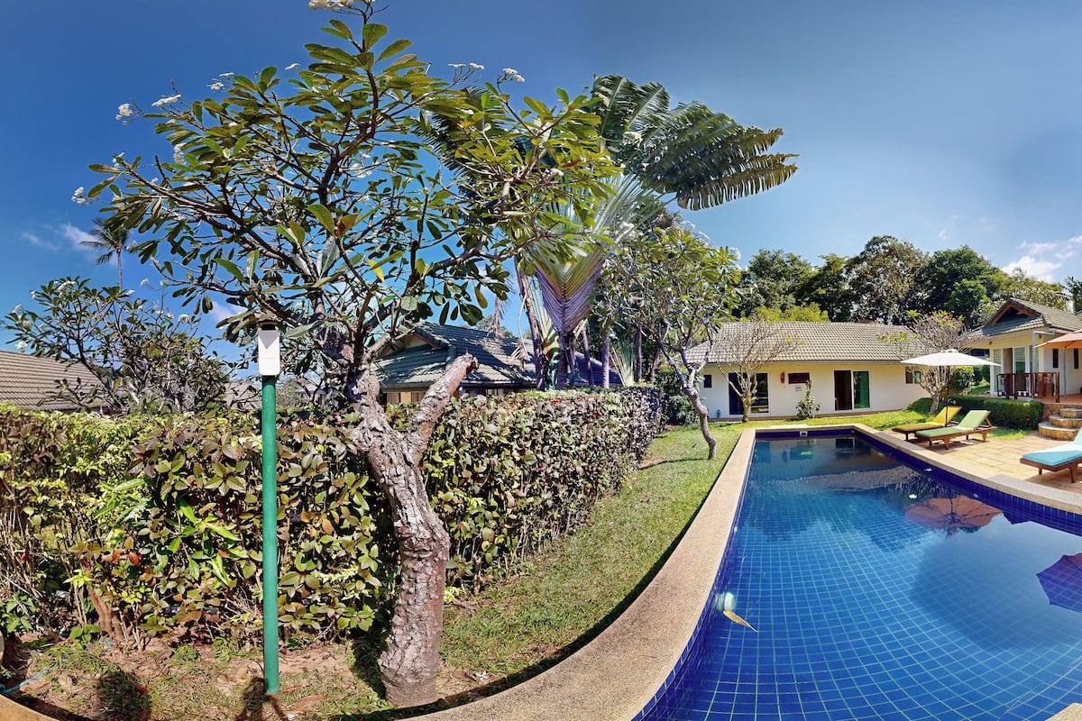 A swimming pool with lounge chairs, surrounded by a well-maintained garden and a house with a tiled roof.