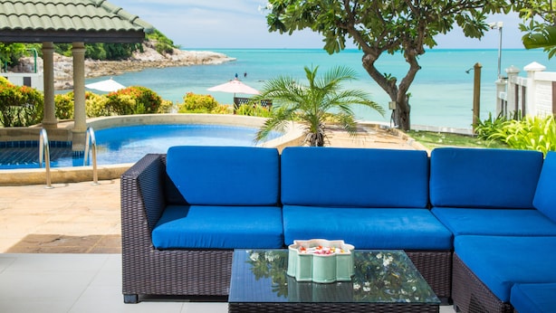 Zona junto a la piscina con un sofá azul y una mesa, con vistas a un mar azul cristalino y una costa rocosa.