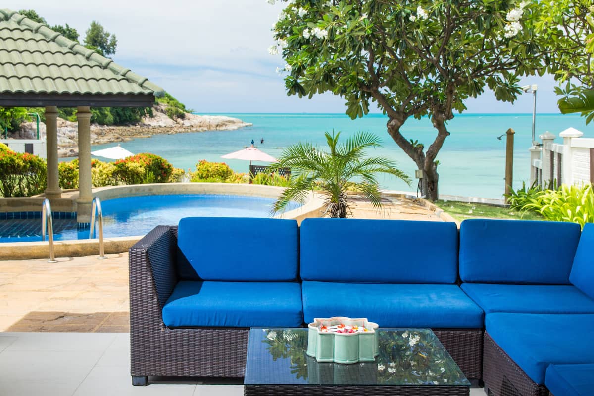 A poolside area with a blue sofa and a table, overlooking a clear blue sea and a rocky coastline.