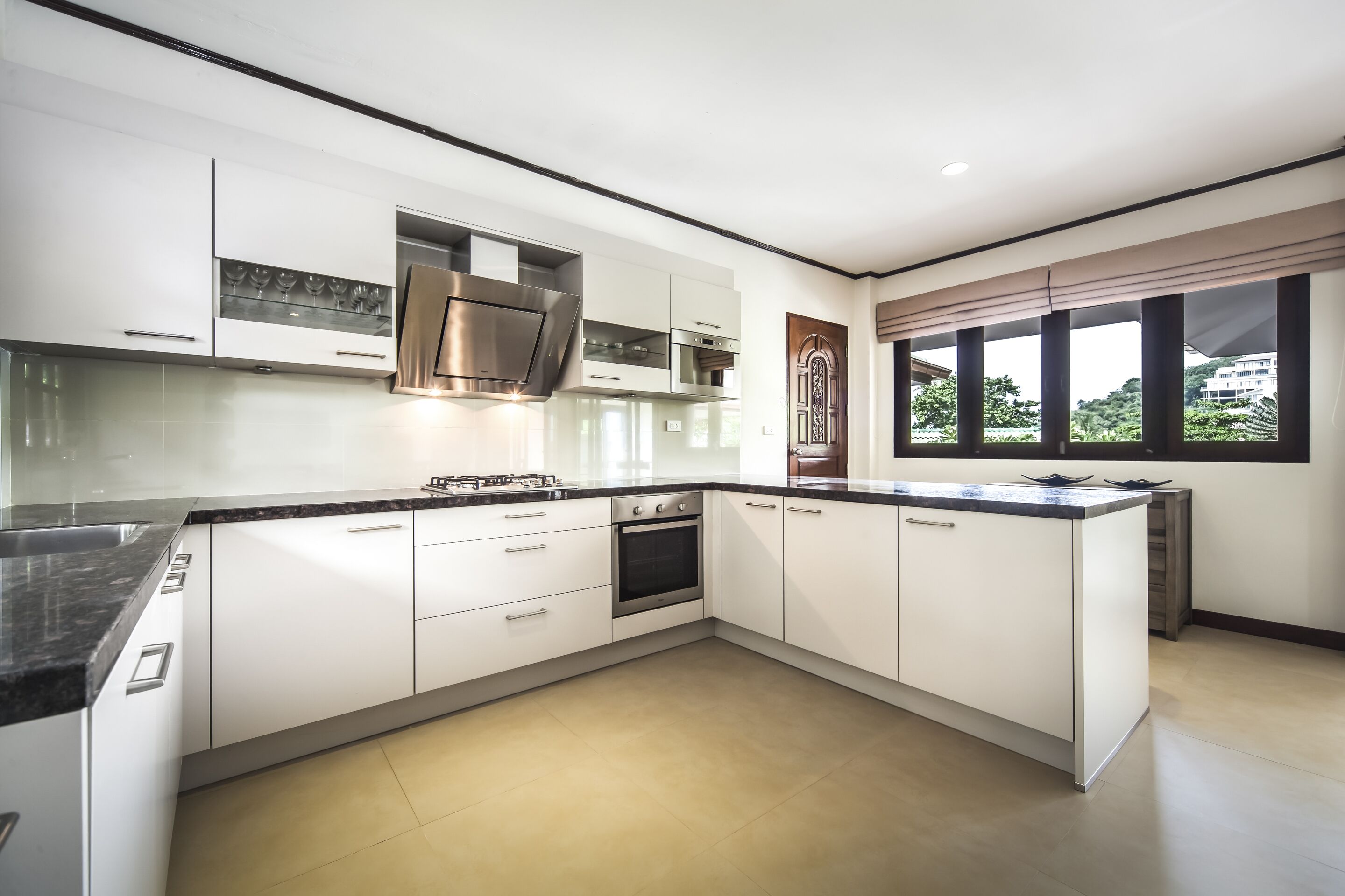 A modern kitchen with white cabinets, a stainless steel range hood, and a large window with a view of greenery.