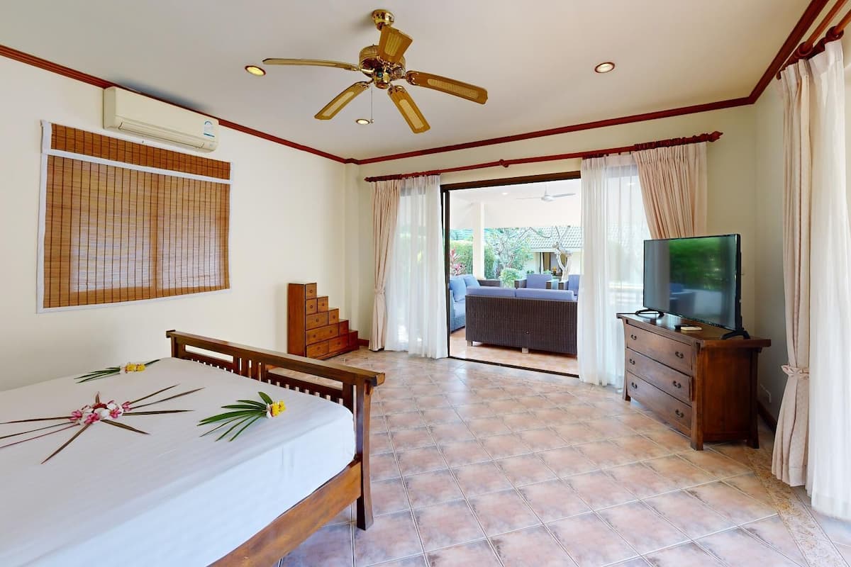 A hotel room with a bed, a TV, a wooden dresser, and a view of an outdoor area with a pool.