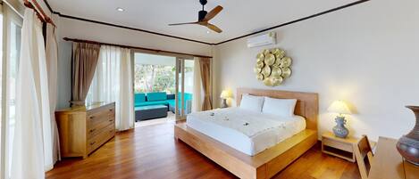 A bedroom with a wooden bed, a ceiling fan, an air conditioning unit, a nightstand with a lamp, and a view of a balcony with a sofa.