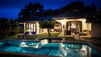 A swimming pool with a tiled edge, a lounge area with chairs and a table, and a building with a tiled roof and glass doors.