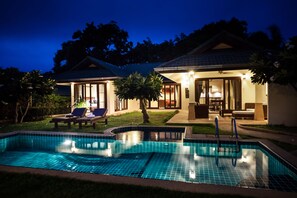 A swimming pool with a tiled edge, a lounge area with chairs and a table, and a building with a tiled roof and glass doors.