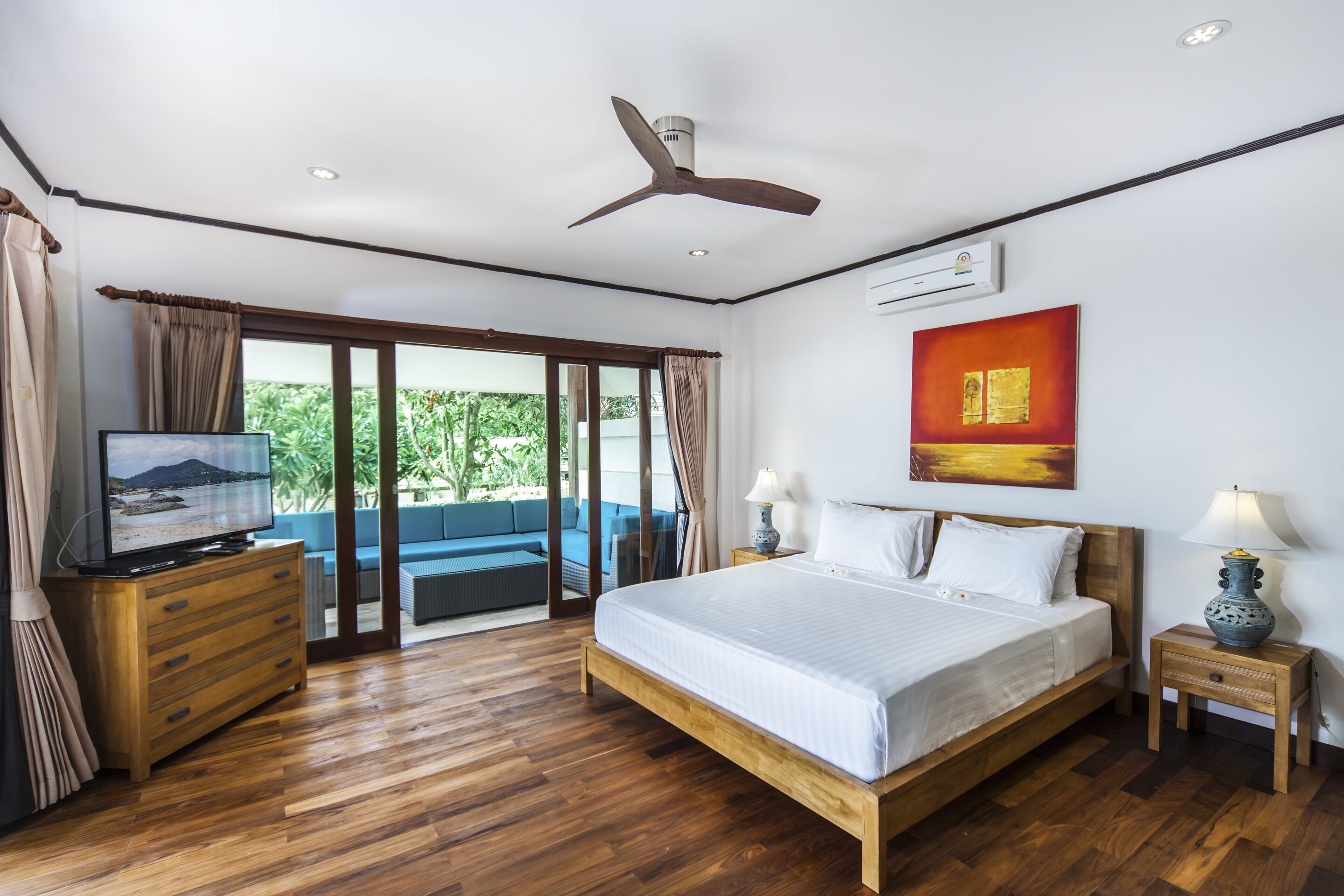 Un dormitorio con cama, piso de madera, ventilador de techo, televisor y vistas a zonas verdes.