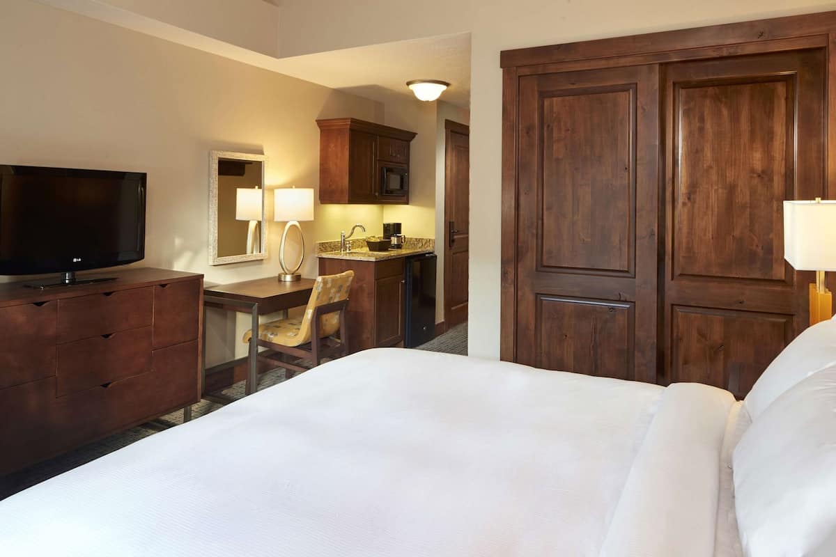 A hotel room with a bed, a TV, a desk with a chair, a mirror, and wooden doors.