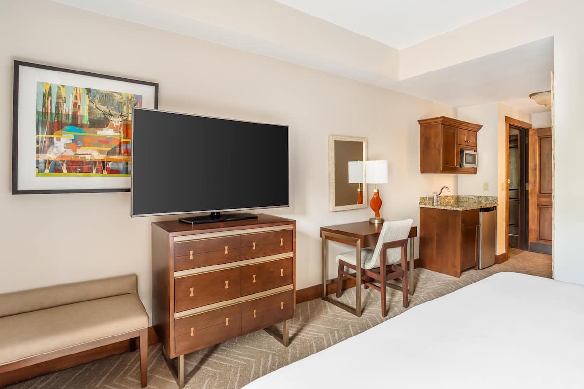 A hotel room with a bed, a TV, a desk with a chair, a small kitchenette, and a painting on the wall.