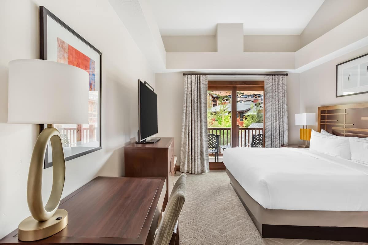 A hotel room with a bed, a desk with a lamp, a TV, and a balcony with curtains.
