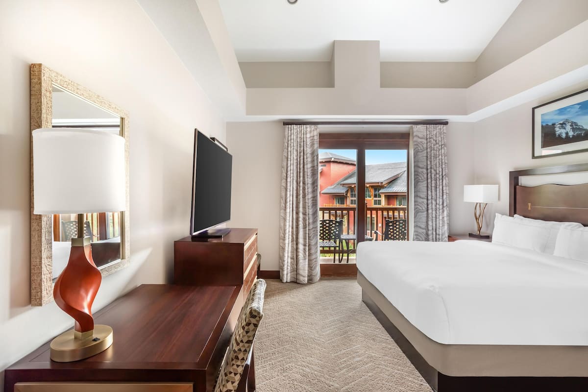 A hotel room with a large bed, a desk with a television, a chair, a desk lamp, and a view of the outside balcony.