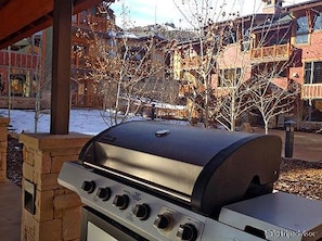 A barbecue grill with four knobs, set against a backdrop of a building with a balcony and leafless trees.