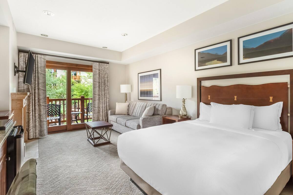 A hotel room with a large bed, a sofa, a small table, and a balcony with curtains.