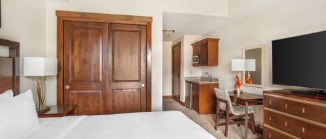 A hotel room with a large bed, a desk, a chair, a television, and a kitchenette.