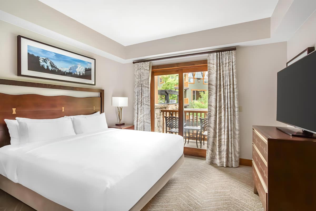 A hotel room with a large bed, a flat-screen TV, a painting on the wall, and a balcony with curtains.