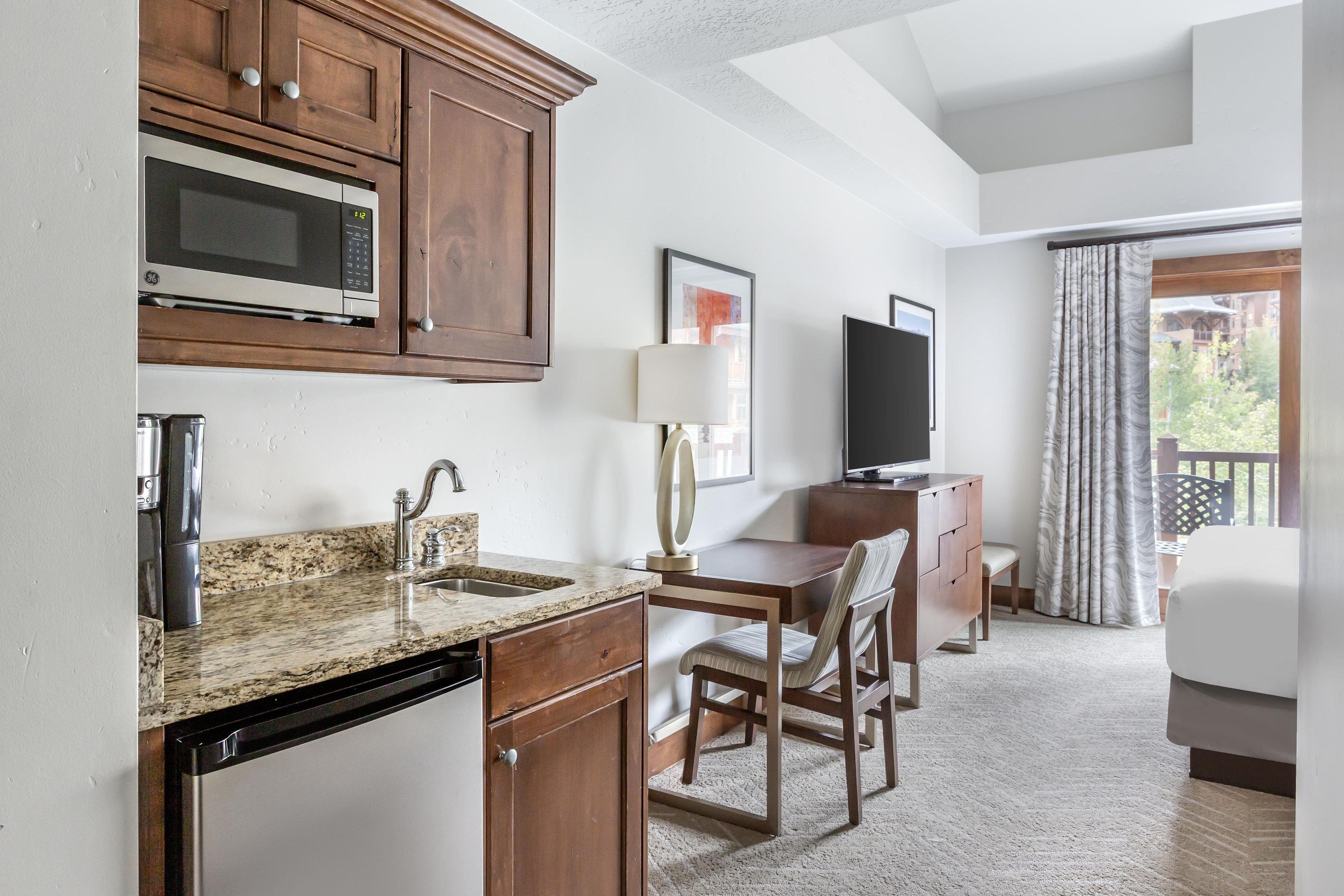 A hotel room with a kitchenette, a bed, a TV, and a window with curtains.